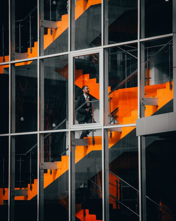 hero-img A businessman in a suit walking on a striking orange staircase in a modern office building.