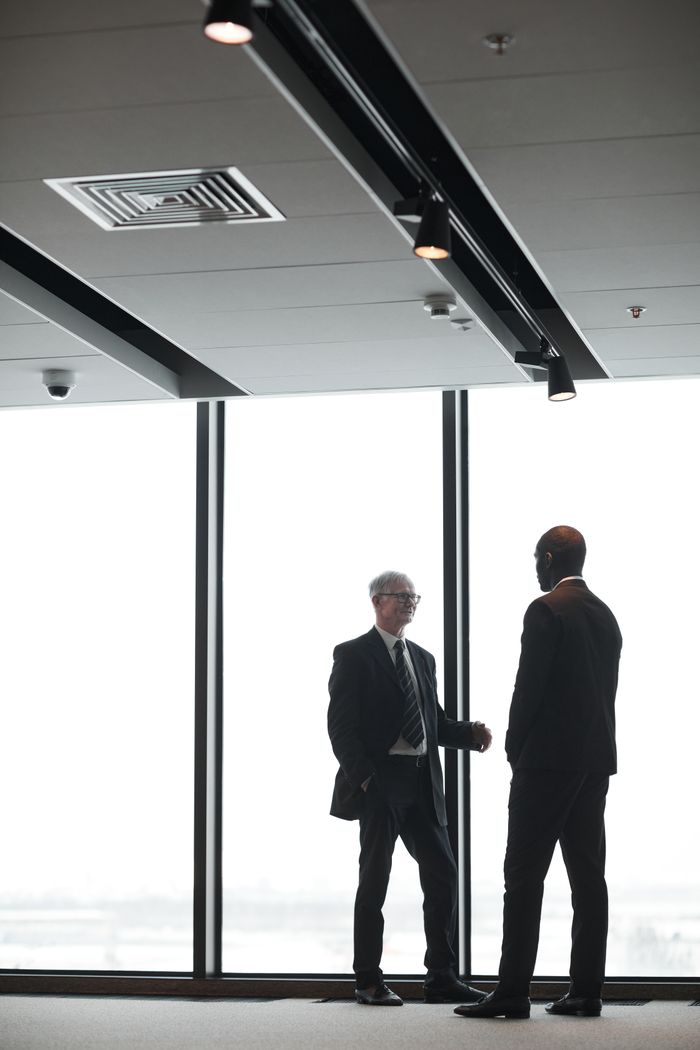 Two corporate executives in formal attire discuss business indoors, silhouetted against large windows.